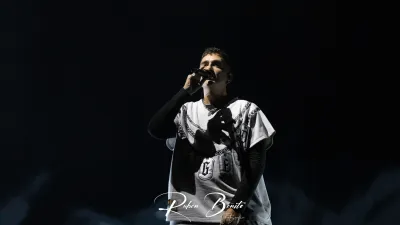 Un miembro de Hijos de la Ruina canta micrófono en mano, con camiseta blanca y gris estampada, en un escenario a oscuras