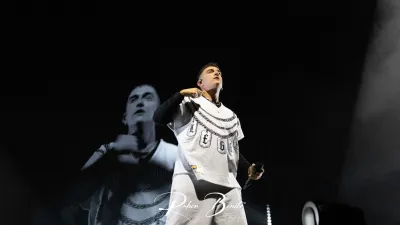 Un integrante de Hijos de la Ruina canta mirando hacia arriba, con camiseta blanca de cadenas, ante su imagen en pantalla
