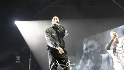 Rapero con chaqueta negra canta con micrófono bajo un foco; a su derecha, otro cantante con jersey blanco y una pantalla al fondo
