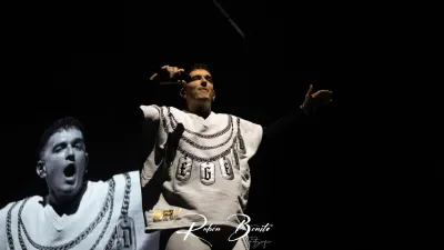 Rapero de Hijos de la Ruina con sudadera blanca de cadenas canta con el brazo extendido, y su imagen ampliada en la pantalla del fondo