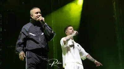 Dos integrantes de Hijos de la Ruina cantan con micrófono, uno de negro y otro de blanco, bajo un foco verde